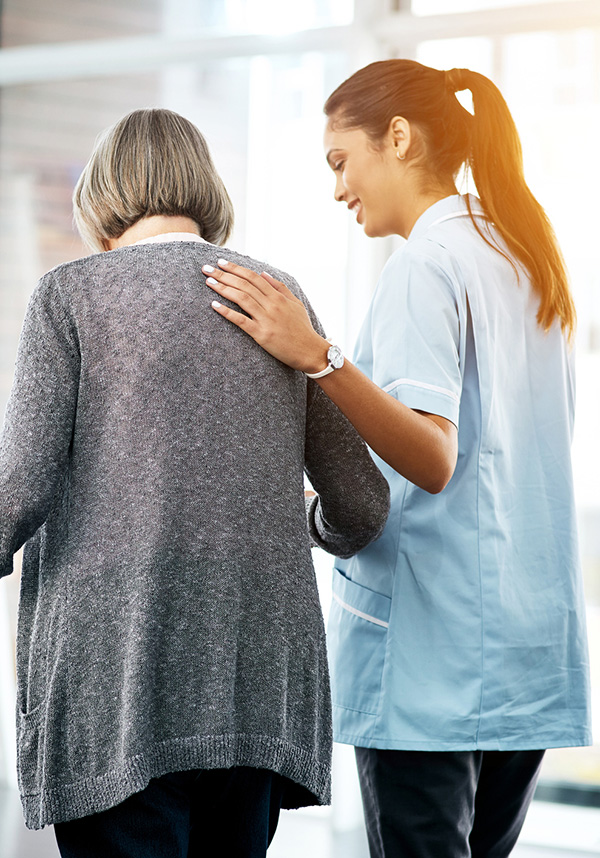 A nurse helps an elderly person walk by putting their hand on the patient's back to guide them.